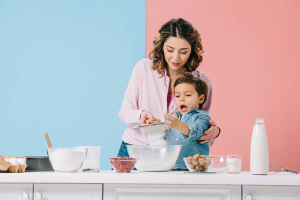 mother with amused little son sifting flour to bowl on bicolor background