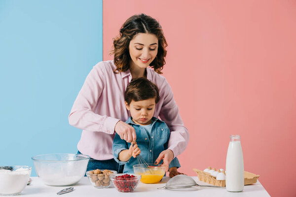 smiling mother helping little son in whipping eggs with balloon whisk on bicolor background