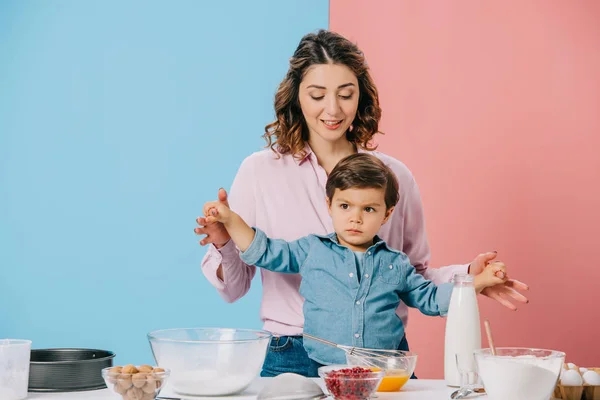 happy mother holding little son hands while standing together by white kitchen table with cooking product and utensils on bicolor background