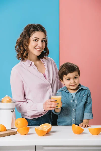 愛らしいに一杯の新鮮なオレンジ ジュースを与えること幸せな母笑顔二色の背景に幼い息子 — ストック写真
