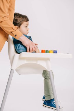 mother hugging little son sitting on highchair with watercolors set on table isolated on white clipart