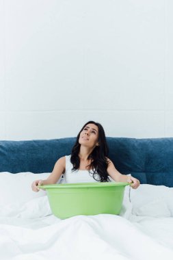 Upset brunette woman sitting on bed and holding basin clipart