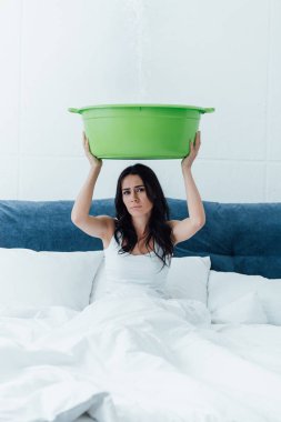 Upset young woman holding basin during leak in bedroom clipart