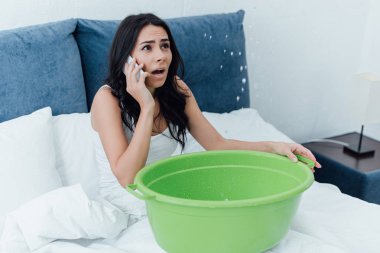 Frightened woman talking on smartphone and looking at water leak in bedroom clipart