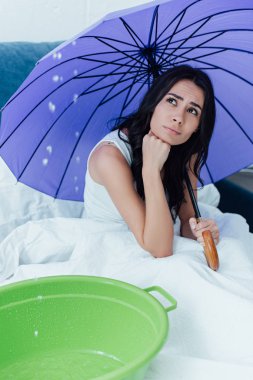 Sad girl with umbrella looking at water leaking from ceiling in bedroom clipart
