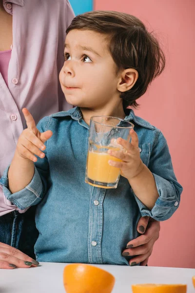 かわいい男の子いっぱい新鮮なオレンジ ジュースのグラスを押しバイカラーの背景に台所のテーブルで立っている母親を見て — ストック写真