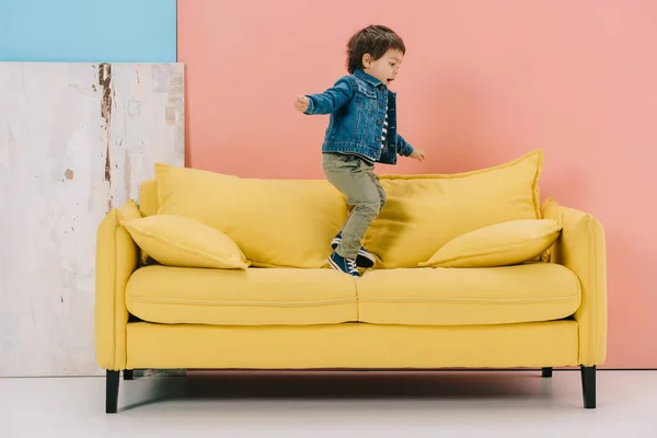 Cute Little Boy Blue Jacket Green Jeans Jumping Yellow Sofa — Stock Photo, Image