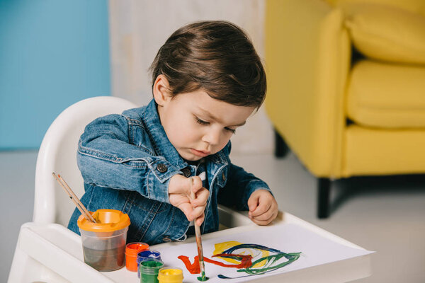 cute little boy painting with watercolor paints while sitting on highchair