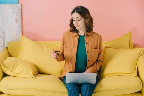 微笑着迷人的女人拿着信用卡 而坐在黄色沙发上 使用笔记本电脑 — 图库照片
