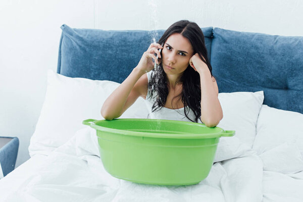 Irritated brunette woman talking on smartphone while sitting in bed with basin