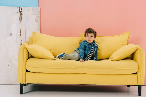 Cute little boy in blue jacket and green jeans sitting on yellow sofa — Stock Image Cute Little Boy Blue Jacket Green Jeans Sitting Yellow Sofa Stock Image