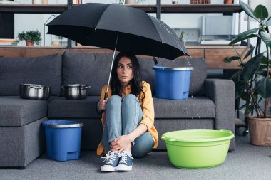 Confused girl sitting under umbrella in living room clipart