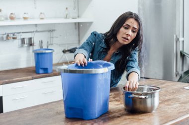 Stressed young woman using buckets and pot during leak in kitchen clipart