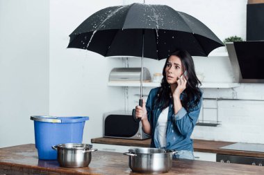 Shocked woman with umbrella dealing with water damage in kitchen and talking on smartphone clipart