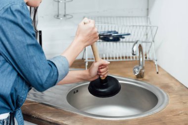 Partial view of woman in denim shirt using plunger in kitchen clipart