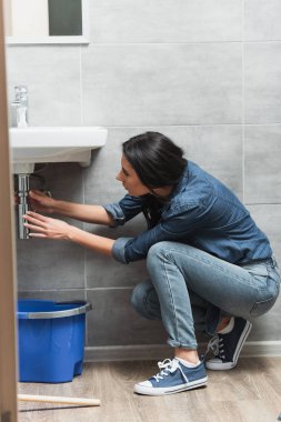 Brunette woman in jeans fixing pipe in bathroom clipart