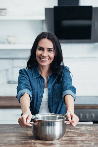 Laughing brunette woman in denim shirt holding pot in kitchen