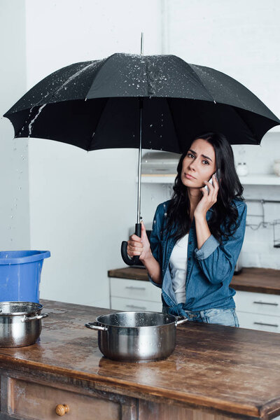 Грустная женщина разговаривает на смартфоне и держит зонтик под протекающим потолком на кухне

