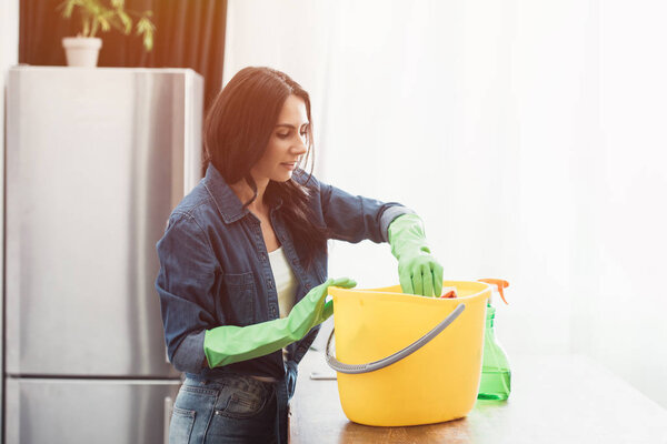 Smiling woman in green rubber gloves using bucket in kitchen