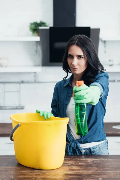 Confident woman in rubber gloves holding spray and bucket in kitchen