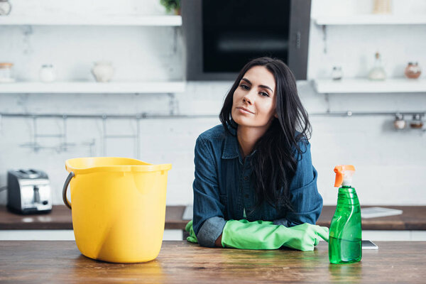 Beautiful brunette woman in rubber gloves sitting with bucket and spray in kitchen