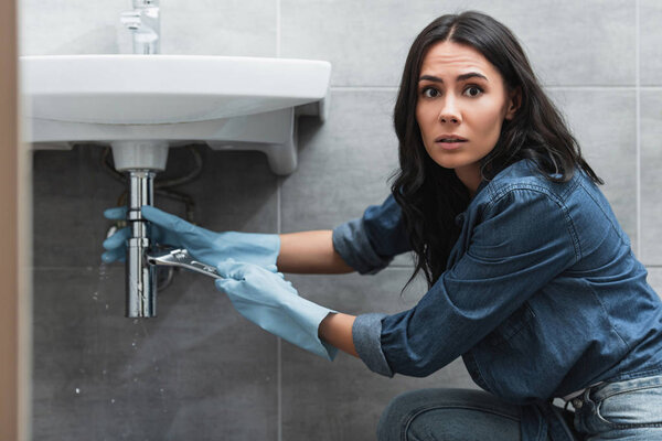 Worried woman in rubber gloves repairing pipe with wrench