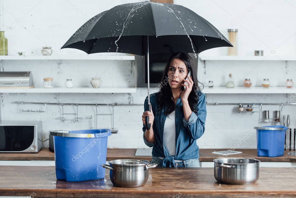 Upset woman standing under umbrella in kitchen and calling plumber