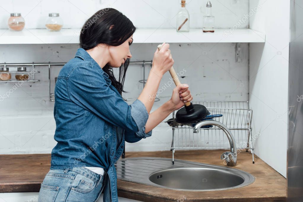 Concentrated woman in denim shirt using plunger in kitchen