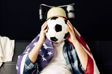 man wearing beer helmet covering face with football and sitting with american flag on shoulders clipart