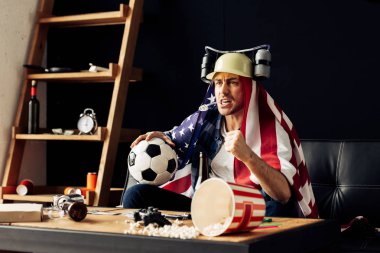 man wearing beer helmet drinking and holding ball while sitting with american flag on shoulders and watching game clipart