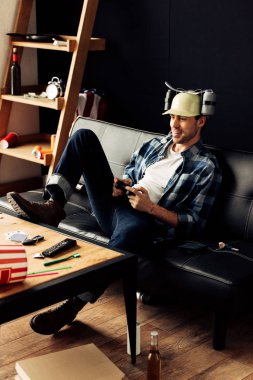 cheerful man in beer helmet playing video game at home clipart