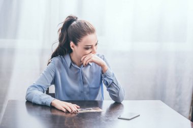 upset woman in blue blouse sitting at table with photo and thinking in room, grieving disorder concept clipart