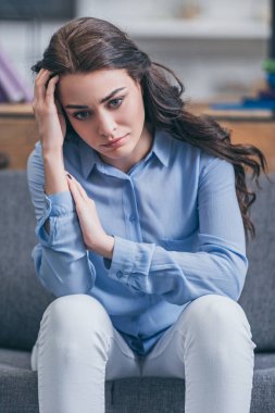 sad woman in blue blouse and white pants sitting on grey couch at home, grieving disorder concept clipart