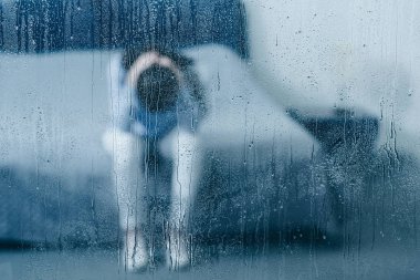 depressed woman sitting on bed and holding head in hands through window with raindrops clipart