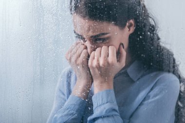 yüzünü elleriyle kapsayan ve yağmur damlaları ile pencereden evde ağlıyor depresif kadın