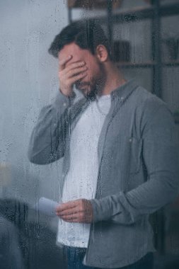 yüz el ile kapsayan, fotoğraf holding ve yağmur damlaları ile pencereden ağlıyor depresif adam