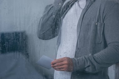 cropped view of man holding photograph through window with raindrops clipart