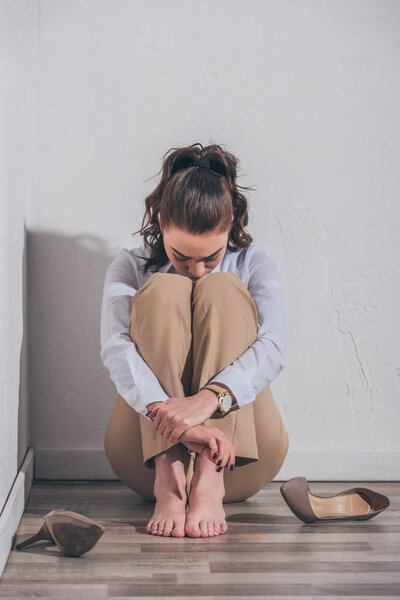 sad woman in white blouse and beige pants sitting on floor, hugging knees near wall at home, grieving disorder concept