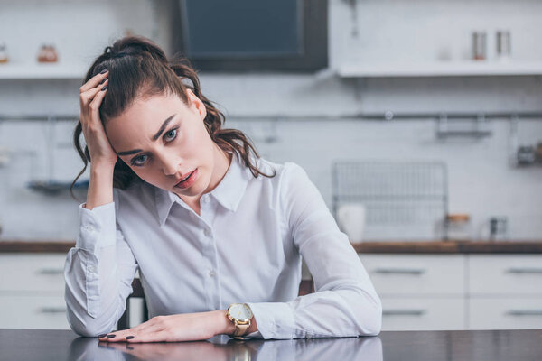 sad woman in white blouse sitting at table in kitchen, grieving disorder concept