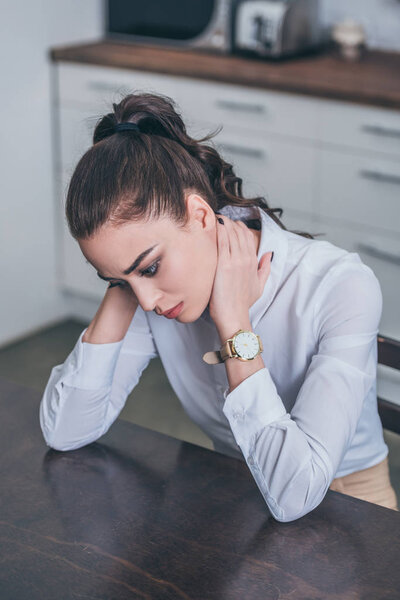 upset woman in white blouse sitting at table in kitchen, thinking and looking down, grieving disorder concept