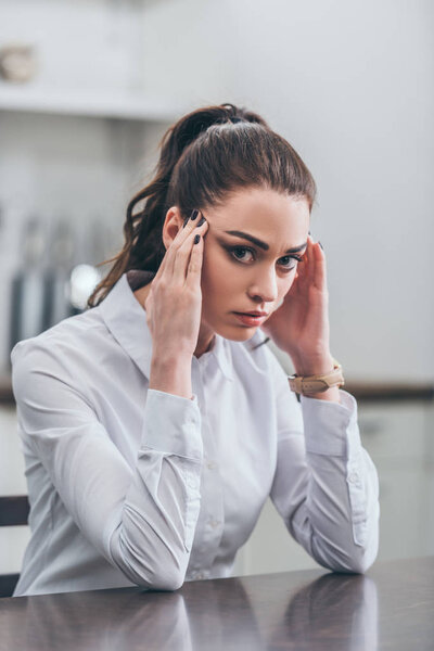 расстроена женщина в белой блузке, сидя за столом на кухне и держа руки на храмах, скорбящее расстройство концепции

