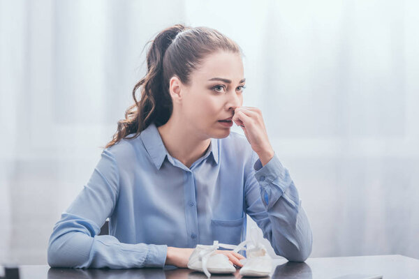 грустная женщина в голубой блузке сидит за деревянным столом в детской обуви, плачет и смотрит вдаль дома, скорбящее расстройство концепции
