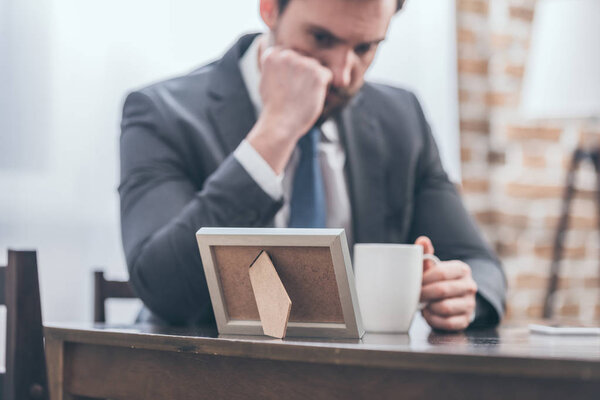 селективное внимание расстроенного мужчины в сером костюме, сидящего за деревянным столом, держащего белую чашку и смотрящего на фоторамку в комнате, концепция расстройства скорби
