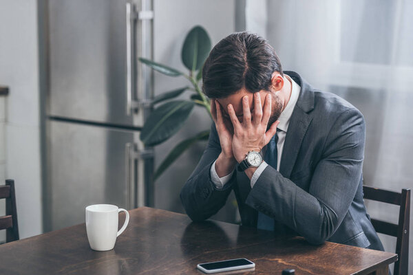 sad man in gray suit sitting at table with smartphone, white cap and covering face with hands at home, grieving disorder concept