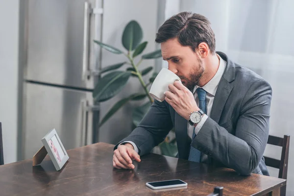 Homem Triste Terno Cinza Sentado Mesa Madeira Com Smartphone Bebendo — Fotografia de Stock