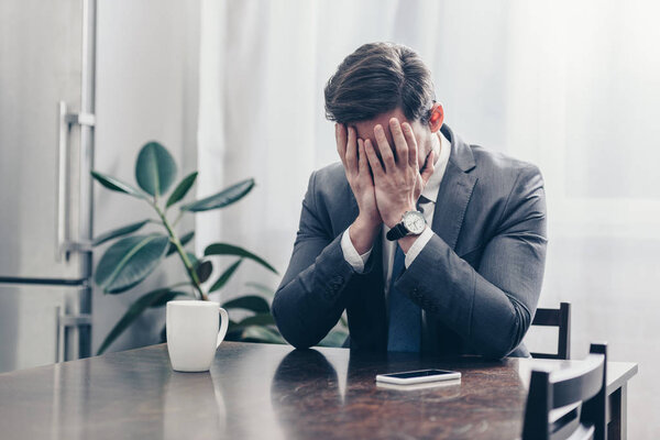 sad man in gray suit sitting at wooden table with smartphone, white cap and covering face with hands at home, grieving disorder concept