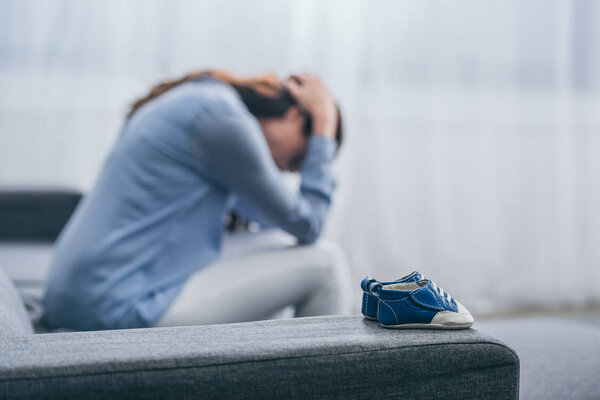 selective focus of blue baby shoes on couch with sad woman sitting and lowering head at home, grieving disorder concept