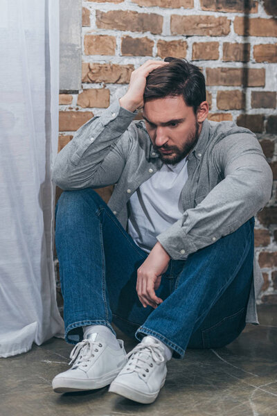 upset man in grey shirt and blue pants sitting on floor on brown textured background in room, grieving disorder concept