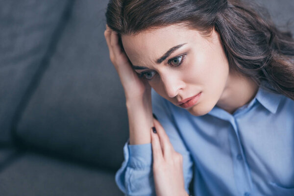sad woman in blue blouse sitting on grey couch and holding head in room, grieving disorder concept