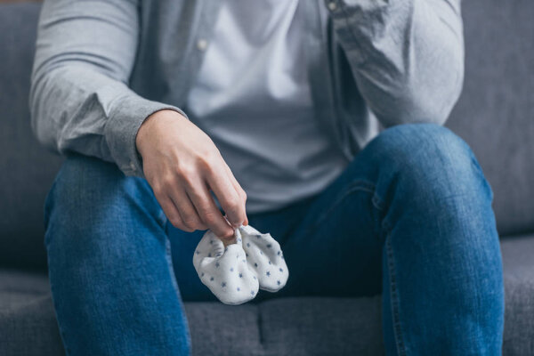 cropped view of man sitting on grey couch and holding babe socks at home, grieving disorder concept